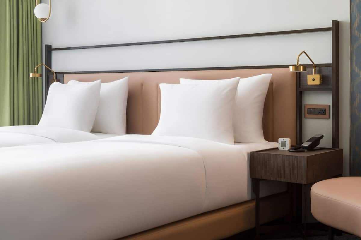 Habitación de hotel con una cama grande, una mesita de noche con un teléfono y una lámpara, una pequeña mesa con una lámpara y una silla.