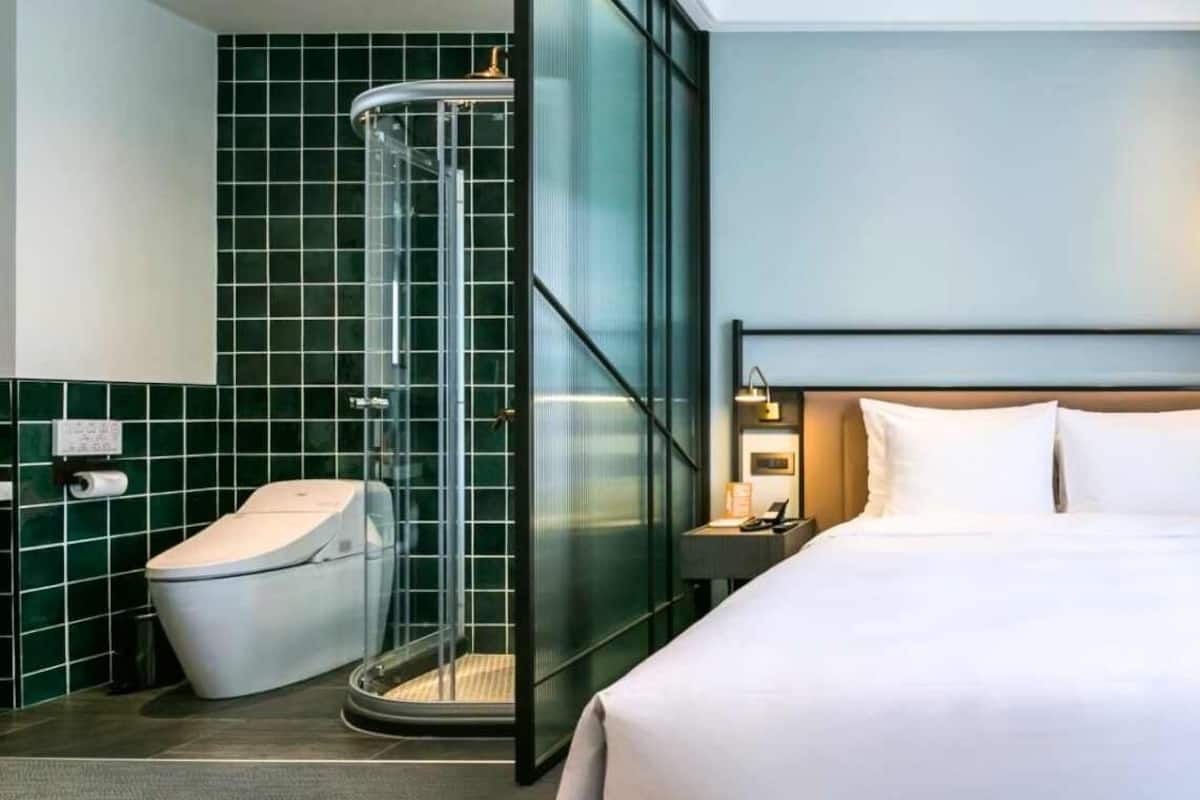 Una habitación de hotel con cama, mesita de noche, lámpara de lectura, ducha con mampara de vidrio y retrete.