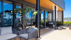 Un espacio exterior moderno con mobiliario, una terraza de madera, ventanas amplias y plantas en macetas.