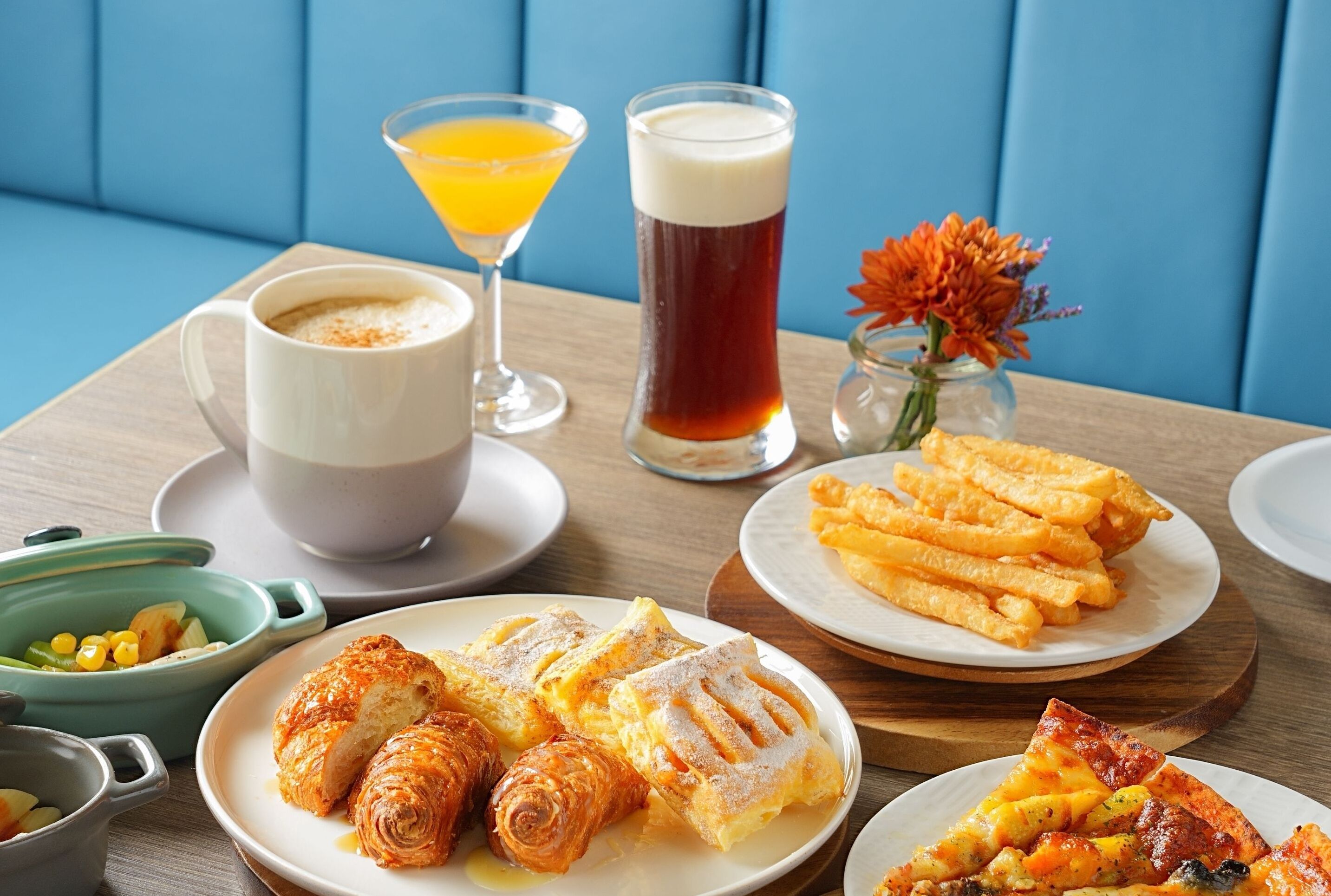 Een ontbijttafel met croissants, een glas sinaasappelsap, een hoog glas bier en een kleine vaas bloemen.