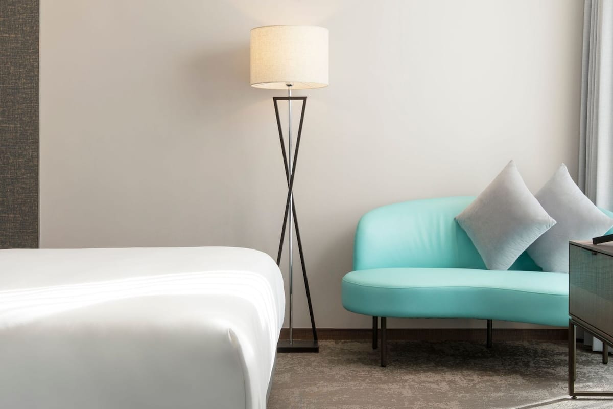 Una habitación de hotel con una cama, un sillón verde azulado, una lámpara de pie y una mesita.