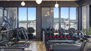 Un gimnasio con equipo de ejercicio, grandes ventanales y vistas a edificios y zonas verdes.