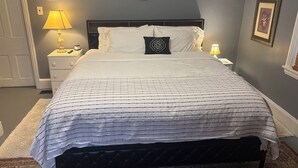 Una cama bien hecha con una colcha a rayas, un espejo redondo encima y una mesita de noche con una lámpara a la izquierda.