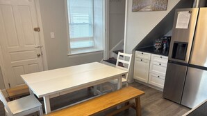 Una cocina con una mesa blanca, un banco de madera, sillas blancas, un refrigerador de acero inoxidable y una ventana con persianas.