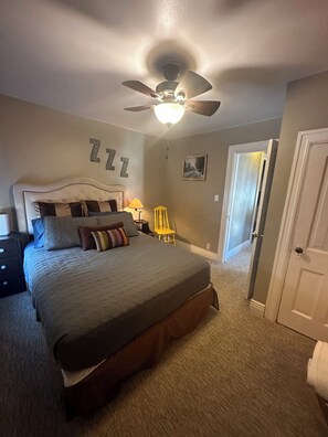 Quarto com cama, ventilador de teto, mesa de cabeceira, luminária, cadeira e porta.