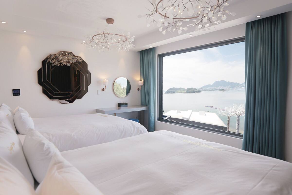 A hotel room with two beds, a large mirror, a chandelier, and a view of a body of water.