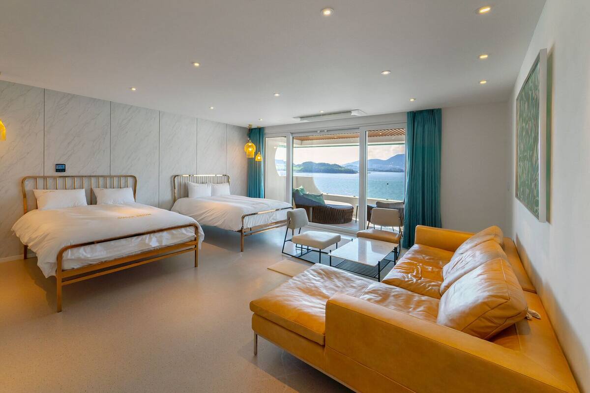 A modern hotel room with two beds, a sofa, a small table, and a view of the sea.