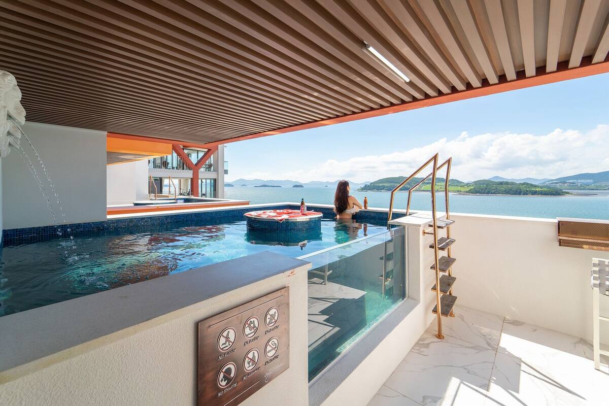 A luxurious outdoor infinity pool with a clear view of the sea and hills, a hot tub, and a wooden pergola.