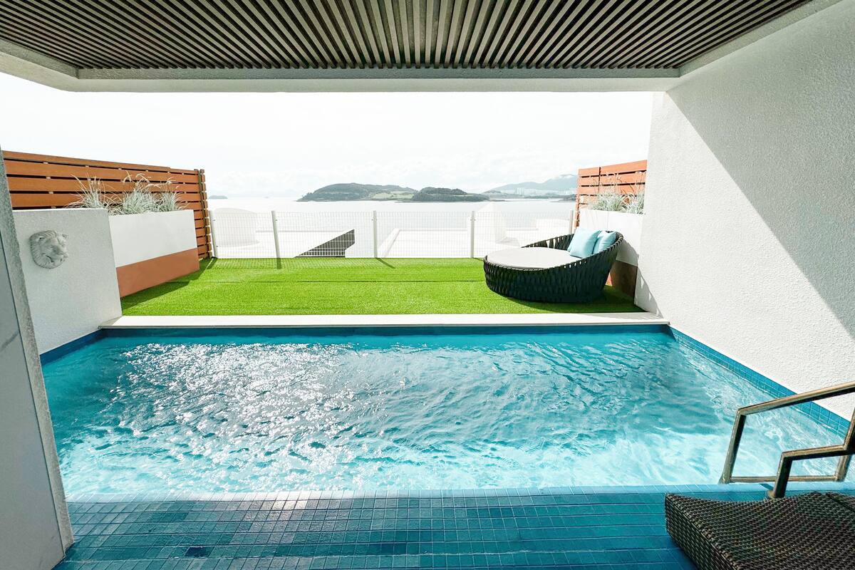A swimming pool with a view of the sea, a wicker lounge chair, and a green lawn area.