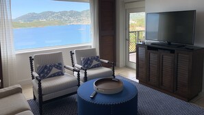 A room with a large window overlooking a body of water and hills, two upholstered chairs with patterned cushions, a round blue ottoman, and a television on a wooden cabinet.