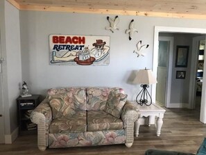 A living room with a floral sofa, a small side table, and a wall-mounted lamp. A beach-themed sign is visible on the wall.