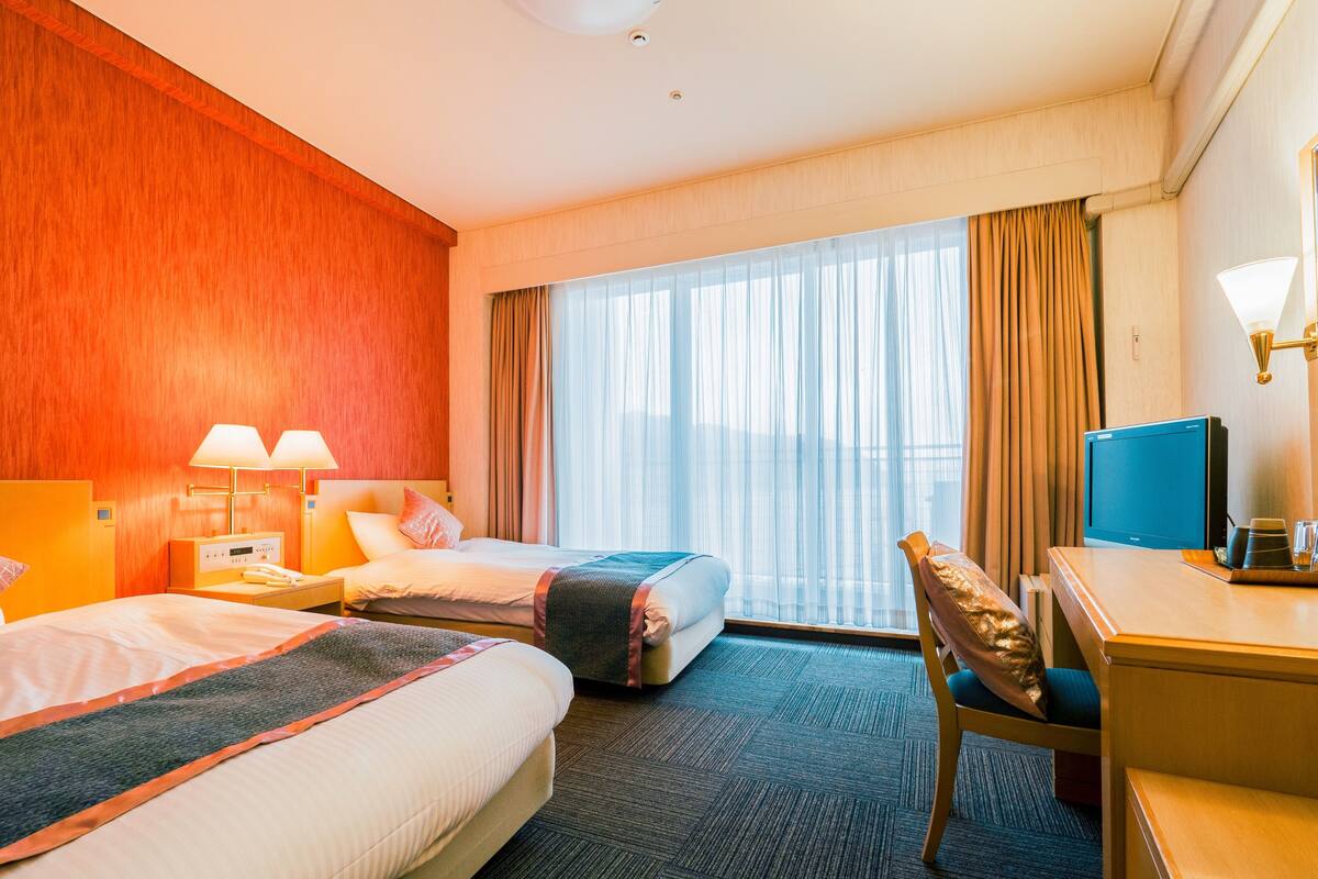 A hotel room with two beds, a desk, a chair, a TV, and a window with curtains.