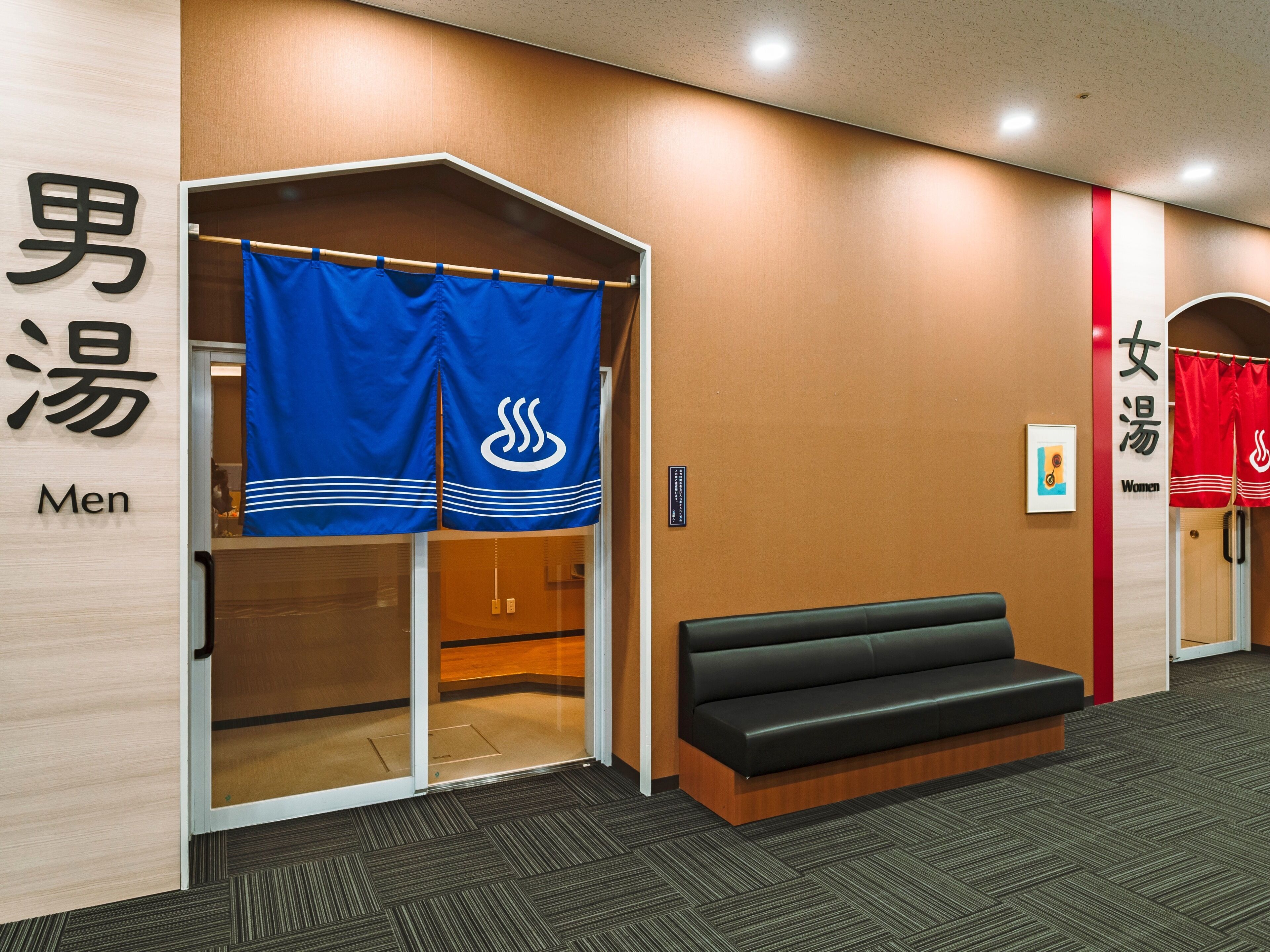 A hotel lobby with a reception desk, a black leather bench, and a wall with signs indicating men's and women's restrooms.