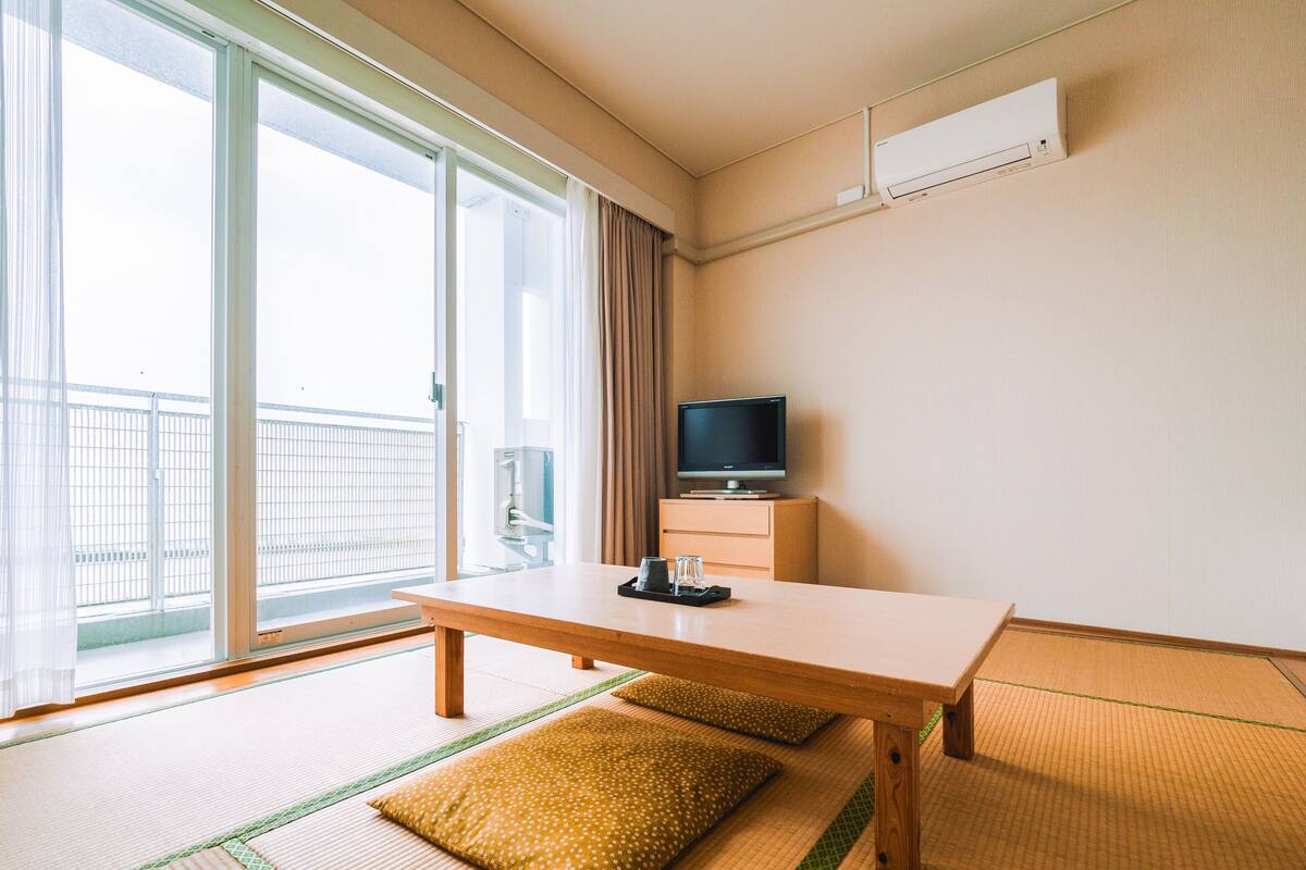 A hotel room with tatami mats, a wooden table, a television on a cabinet, and a sliding glass door to a balcony.