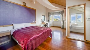 Une chambre avec un grand lit, un plancher en bois, un bureau et une salle de bain visible à travers une porte ouverte.
