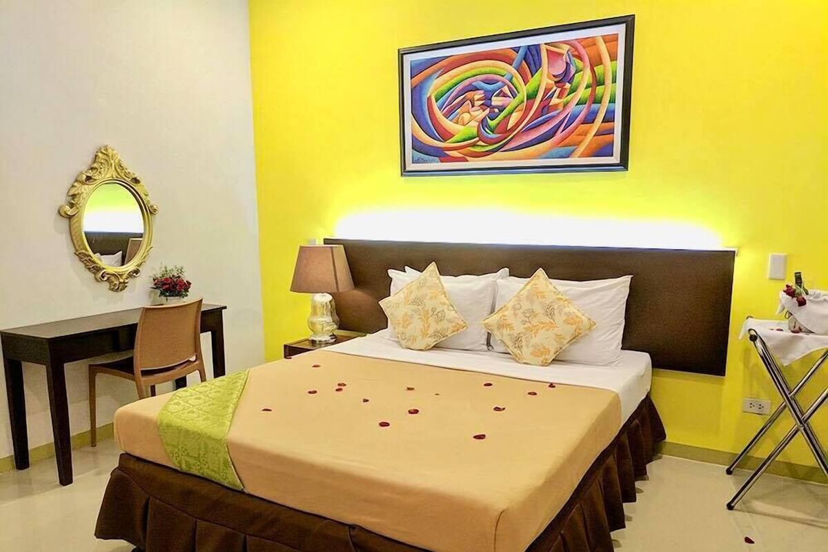 A hotel room with a bed, a desk, a chair, a lamp, and a painting on the wall.