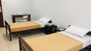 Et hotelværelse med to enkeltsenge, begge med træramme og beige sengetøj. Der er et lille bord med en stol og et natbord med en lampe samt et glas vand.