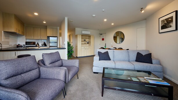 A modern living room with a grey sofa, a glass coffee table, and a kitchen in the background. - Kaikoura Luxury Apartments (Kaikoura)