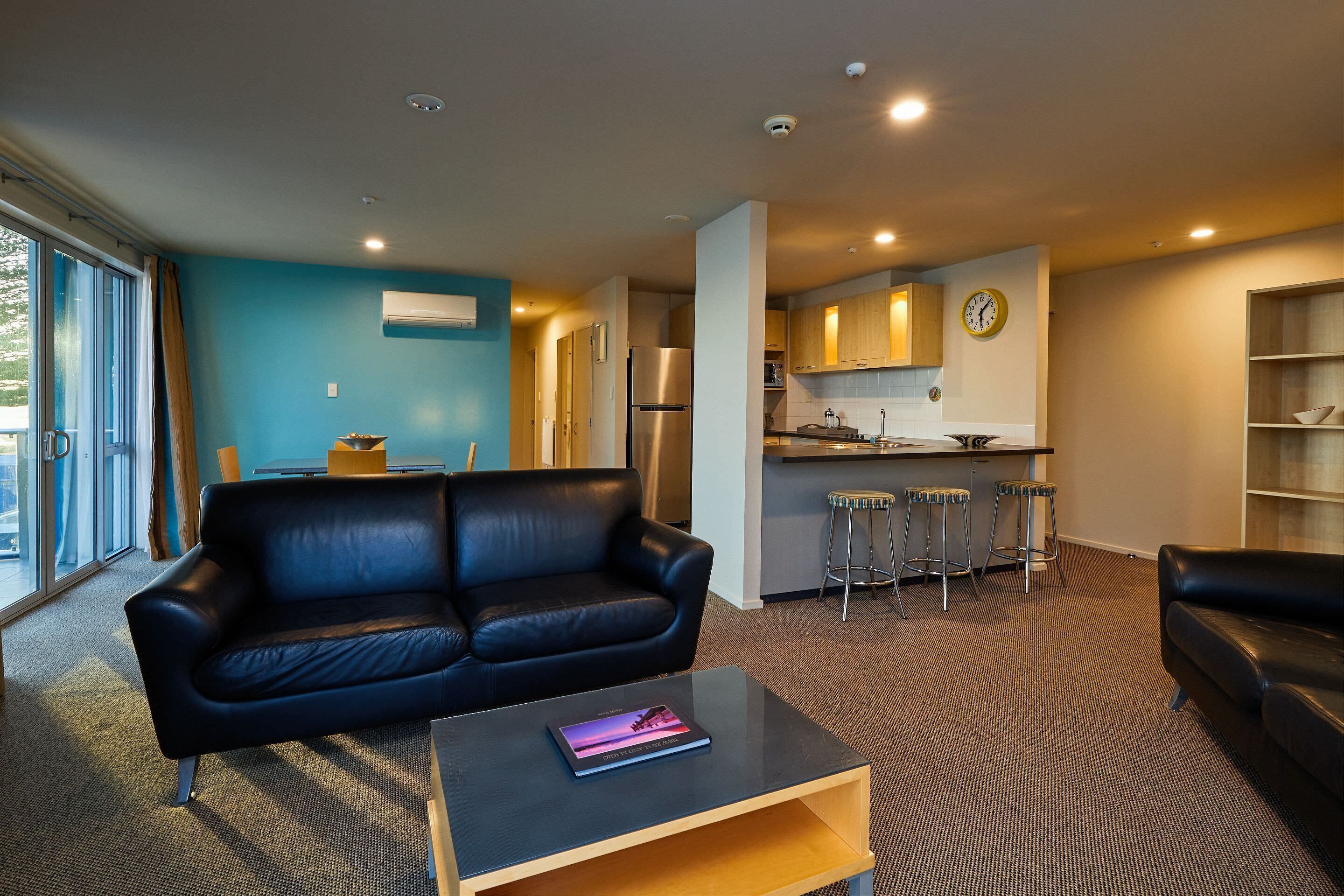 A modern living room with a black leather sofa, a wooden coffee table, and a kitchen area with wooden cabinets and a stainless steel refrigerator.