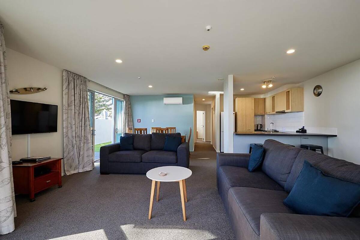 A modern living room with a flat-screen TV, two sofas, a small coffee table, and a kitchen area with wooden cabinets and a sink.