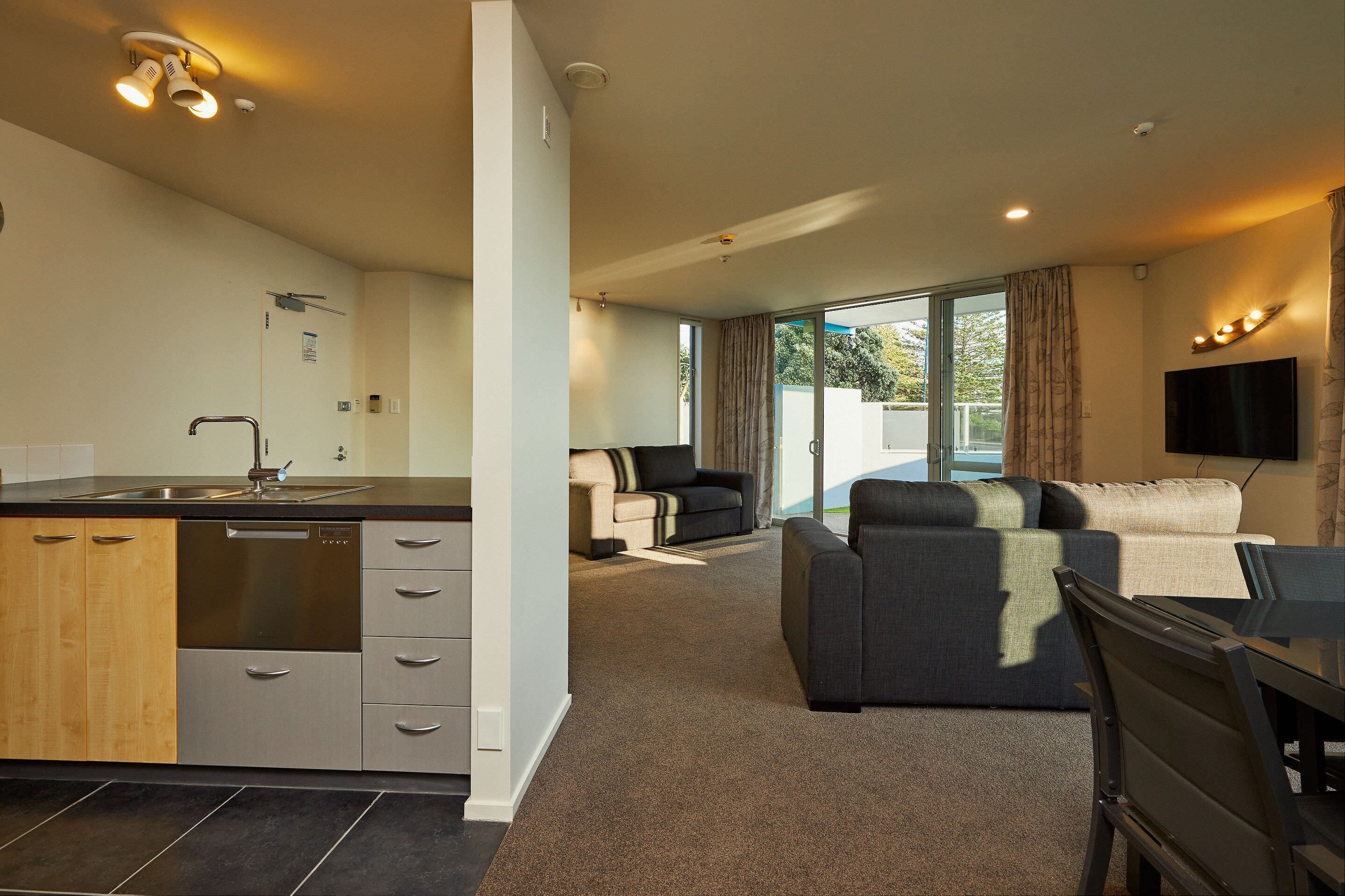 A modern kitchen with a dishwasher, a dining area with a table and chairs, a sofa, and a TV mounted on the wall.