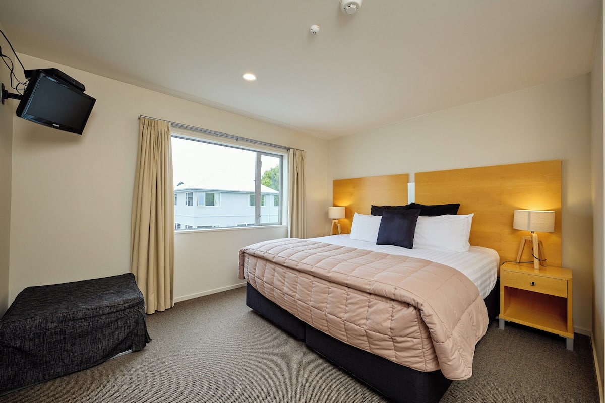 A hotel room with a bed, bedside tables, a TV mounted on the wall, and a window with curtains.
