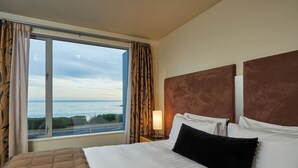 Ein Hotelzimmer mit großem Fenster, einem Bett mit weißen Leinentüchern, einem braunen Kopfteil und einem Nachttisch mit Lampe.