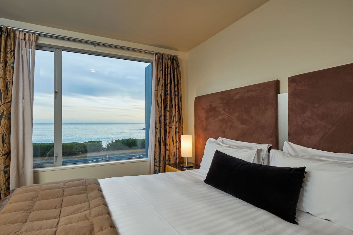 A hotel room with a large window, a bed with white linens, a brown headboard, and a nightstand with a lamp.