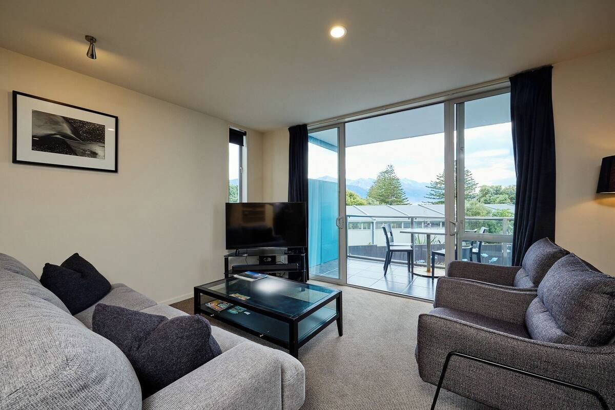A living room with a sofa, a coffee table, a flat-screen TV, and a balcony with a view of mountains.