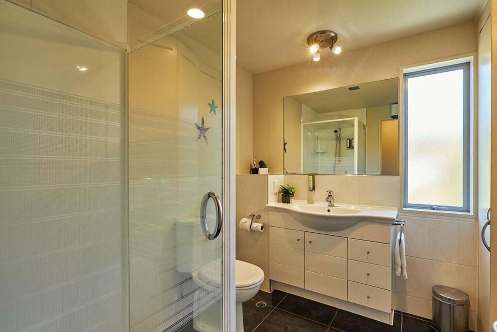 Une salle de bain moderne avec une douche à parois de verre, des toilettes blanches, un lavabo avec un miroir au-dessus et une fenêtre qui laisse entrer la lumière naturelle.