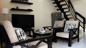 A living room with a flat-screen TV, two armchairs, a coffee table, a staircase, and a shelf with decorative items. - Townhome with 24Hr Security in Panglao Island (Dauis)