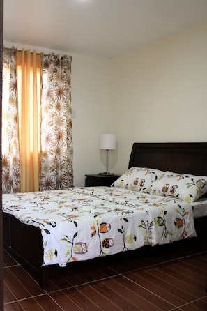 A neatly made bed with a floral-patterned bedspread, a bedside table with a lamp, and curtains with a leaf pattern.