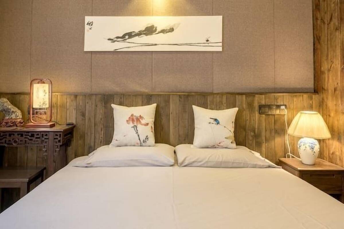 A hotel room with a wooden headboard, two pillows with floral designs, a wall-mounted artwork, a bedside table with a lamp, and a decorative clock.