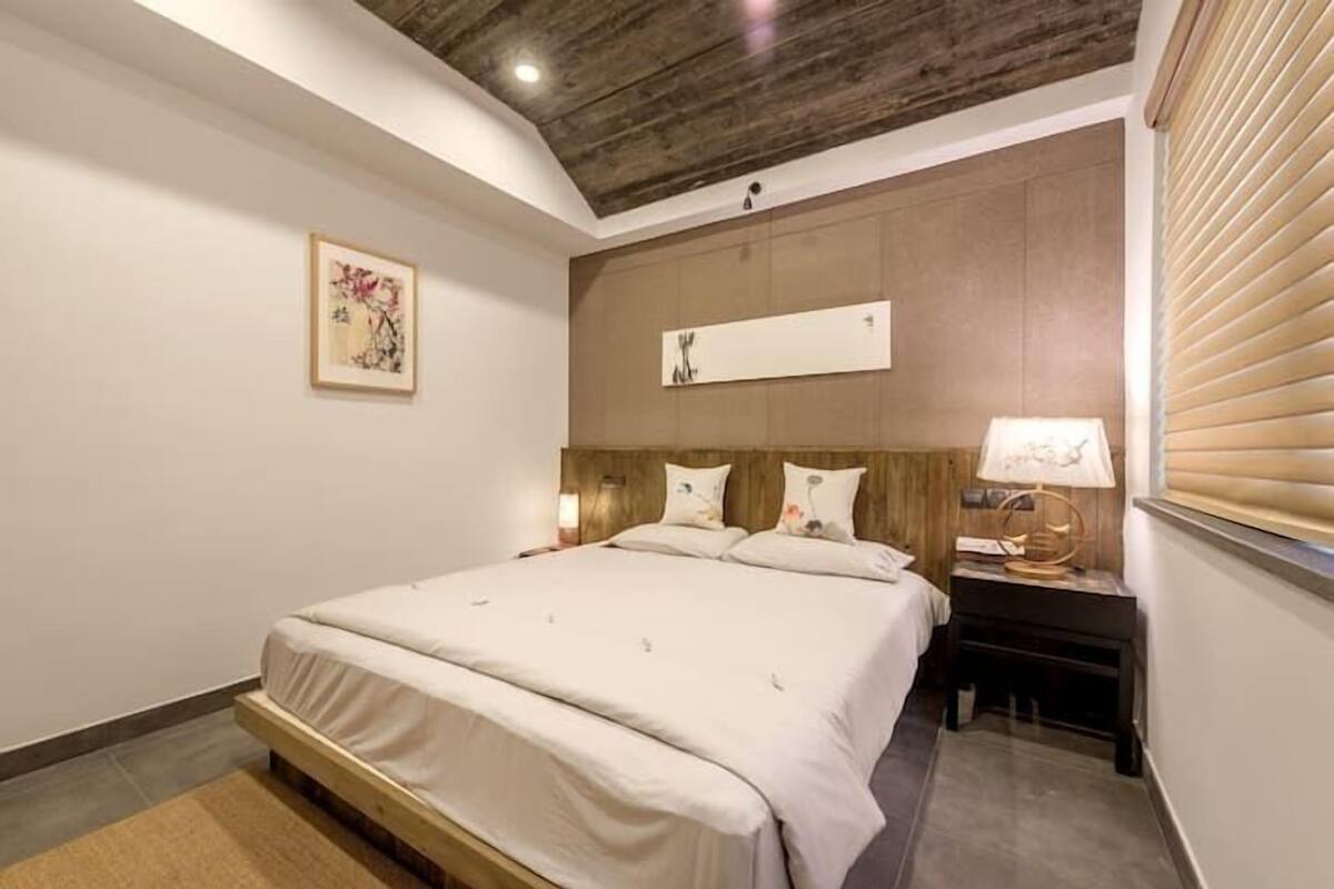 A bedroom with a wooden headboard, a bed with white bedding, a bedside table with a lamp, and a framed picture on the wall.