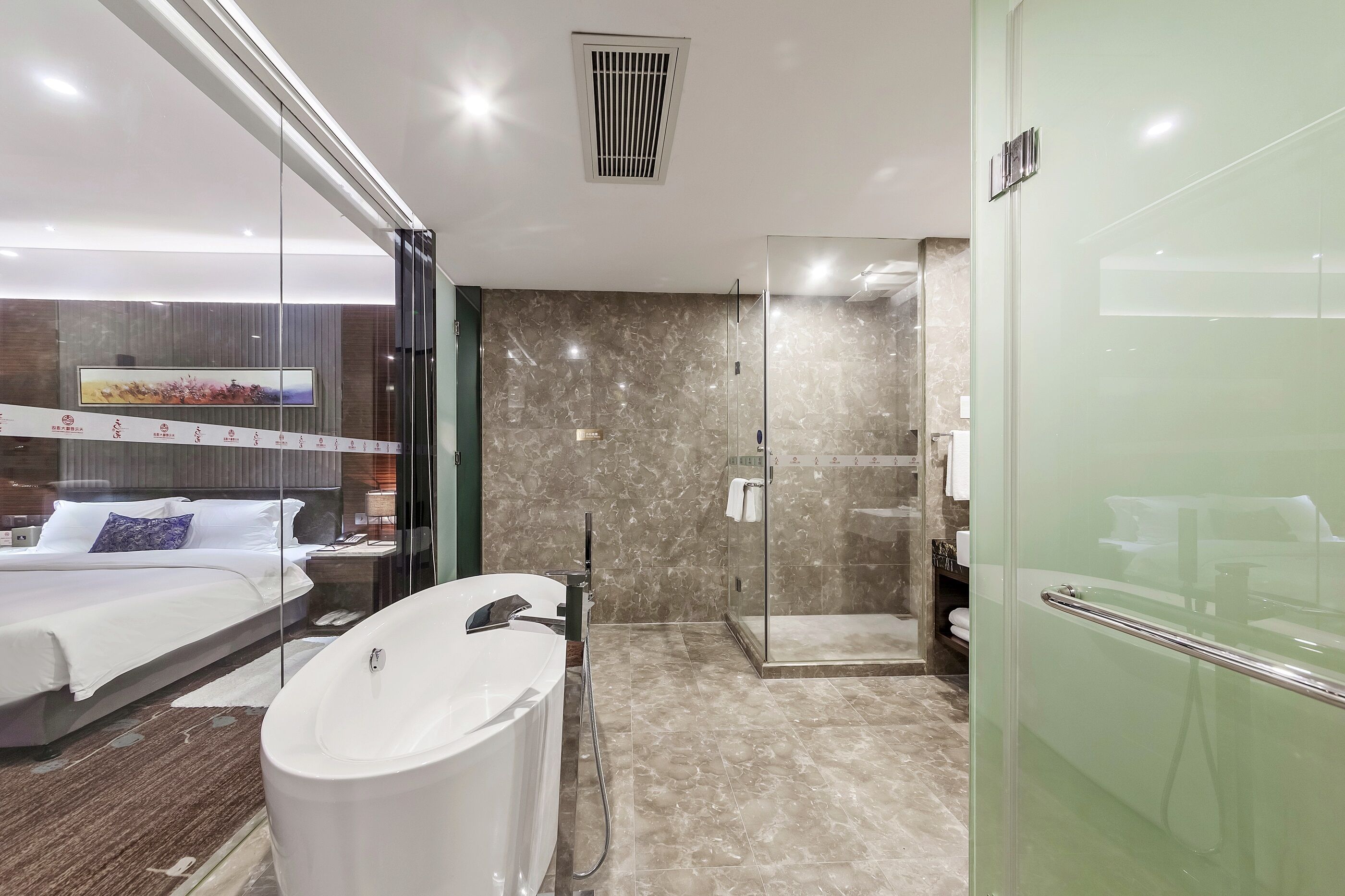 A modern hotel room with a large bed, a bathtub, a glass-enclosed shower, and a large flat-screen TV mounted on the wall.