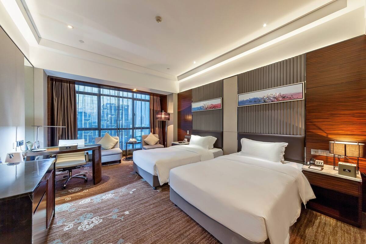 A hotel room with two beds, a desk, a chair, a sofa, a large window, and a city view.