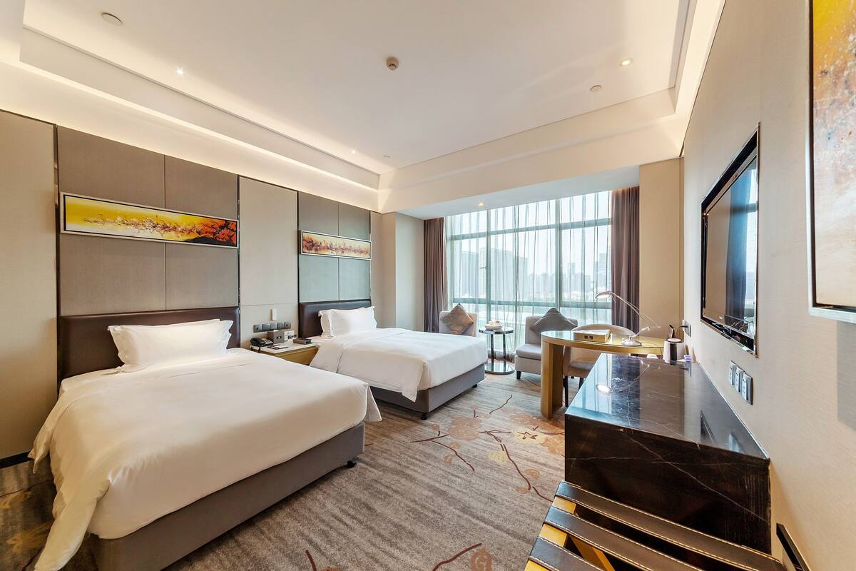 A hotel room with two beds, a desk, a TV, and large windows.