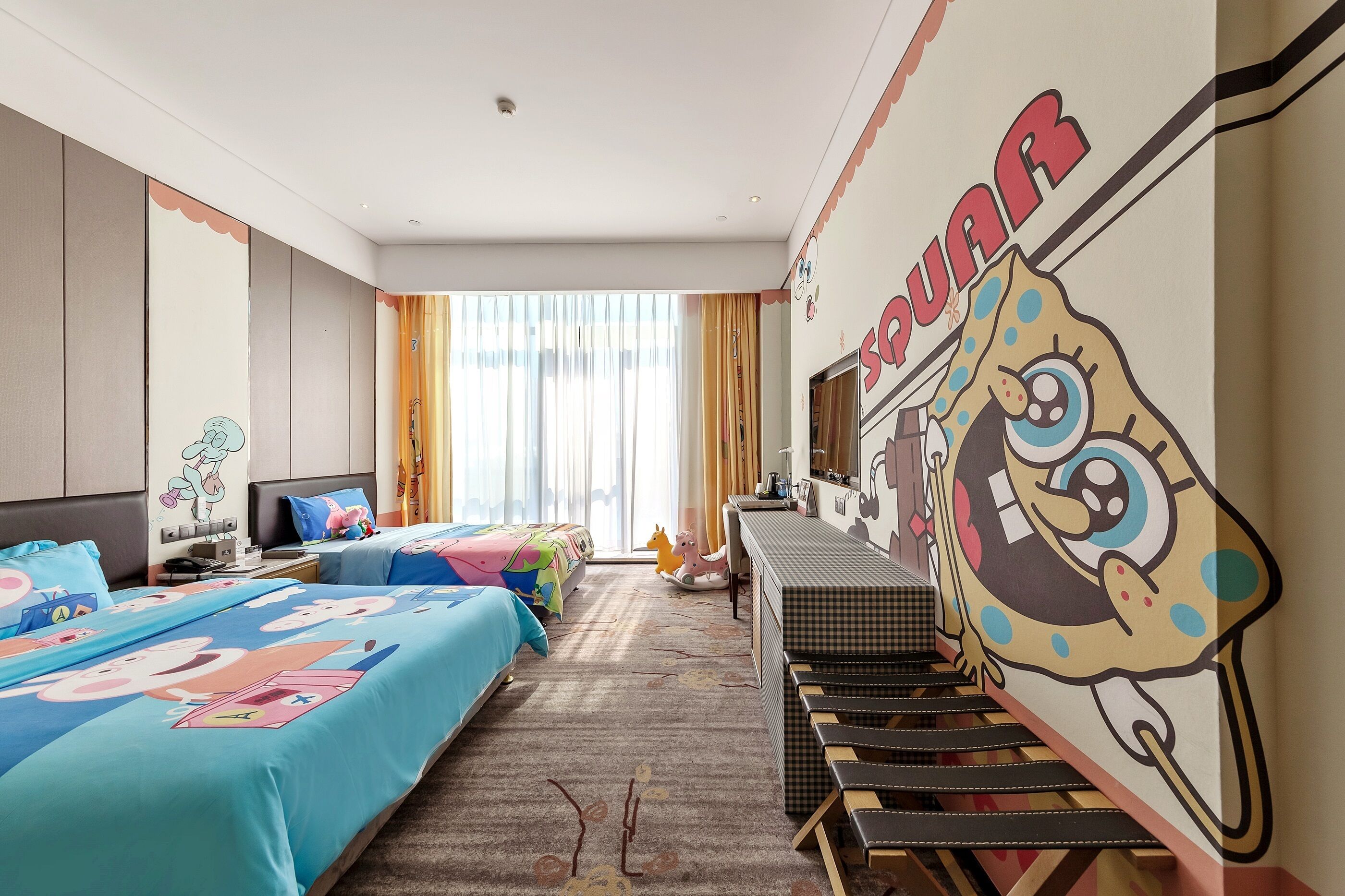 A hotel room with two beds, a large cartoon character mural, a desk, and a window with curtains.