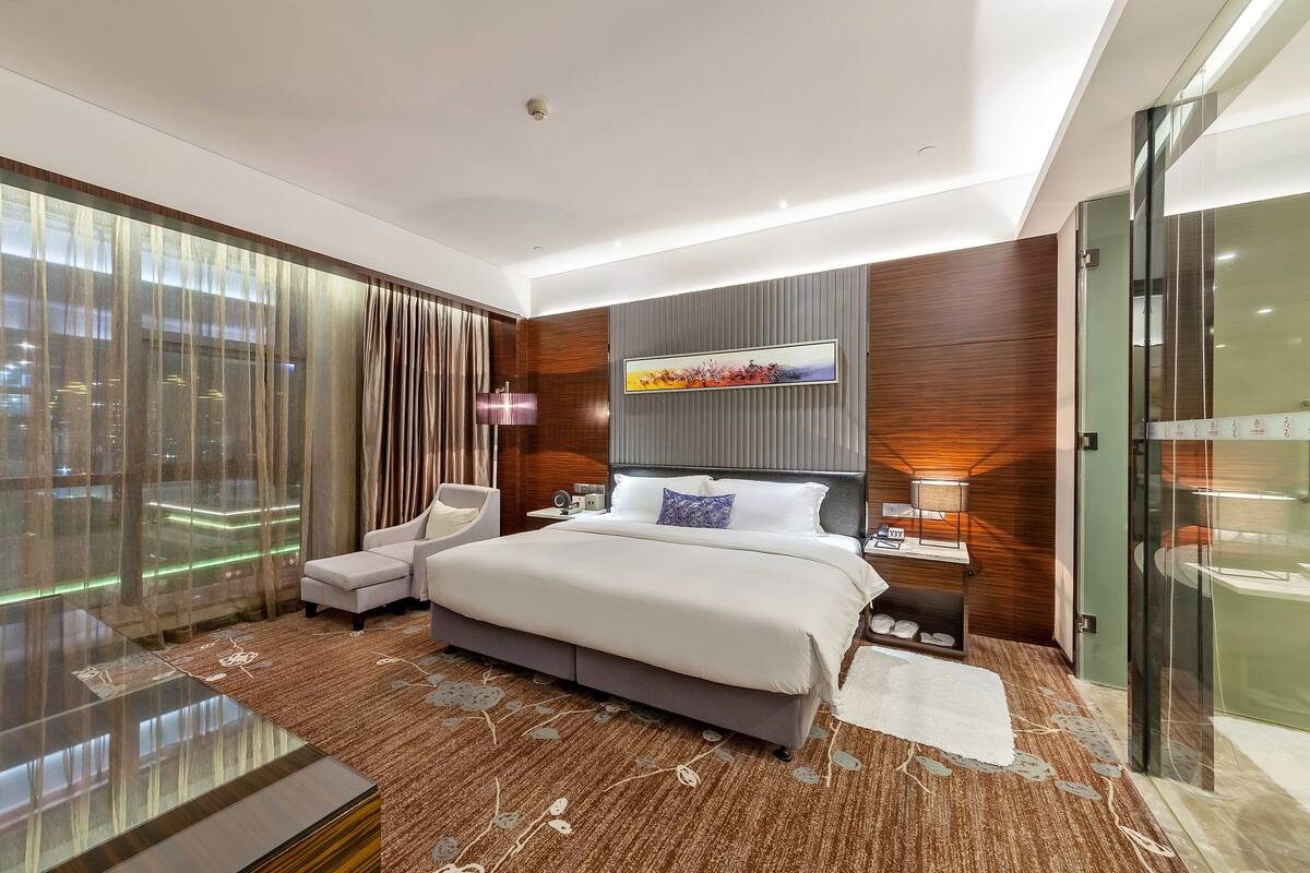 A modern hotel room with a large bed, a glass shower enclosure, and a city view.