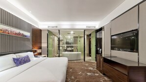 A modern hotel room with a large bed, a flat-screen TV, a bathroom with a glass shower enclosure, and a view of the cityscape. - Tianyuan Junlong Hotel (Xiamen)