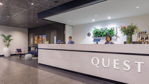A hotel reception area with a modern design, featuring a reception desk labeled "QUEST," a seating area with chairs and a sofa, and a wall adorned with plants and artwork.