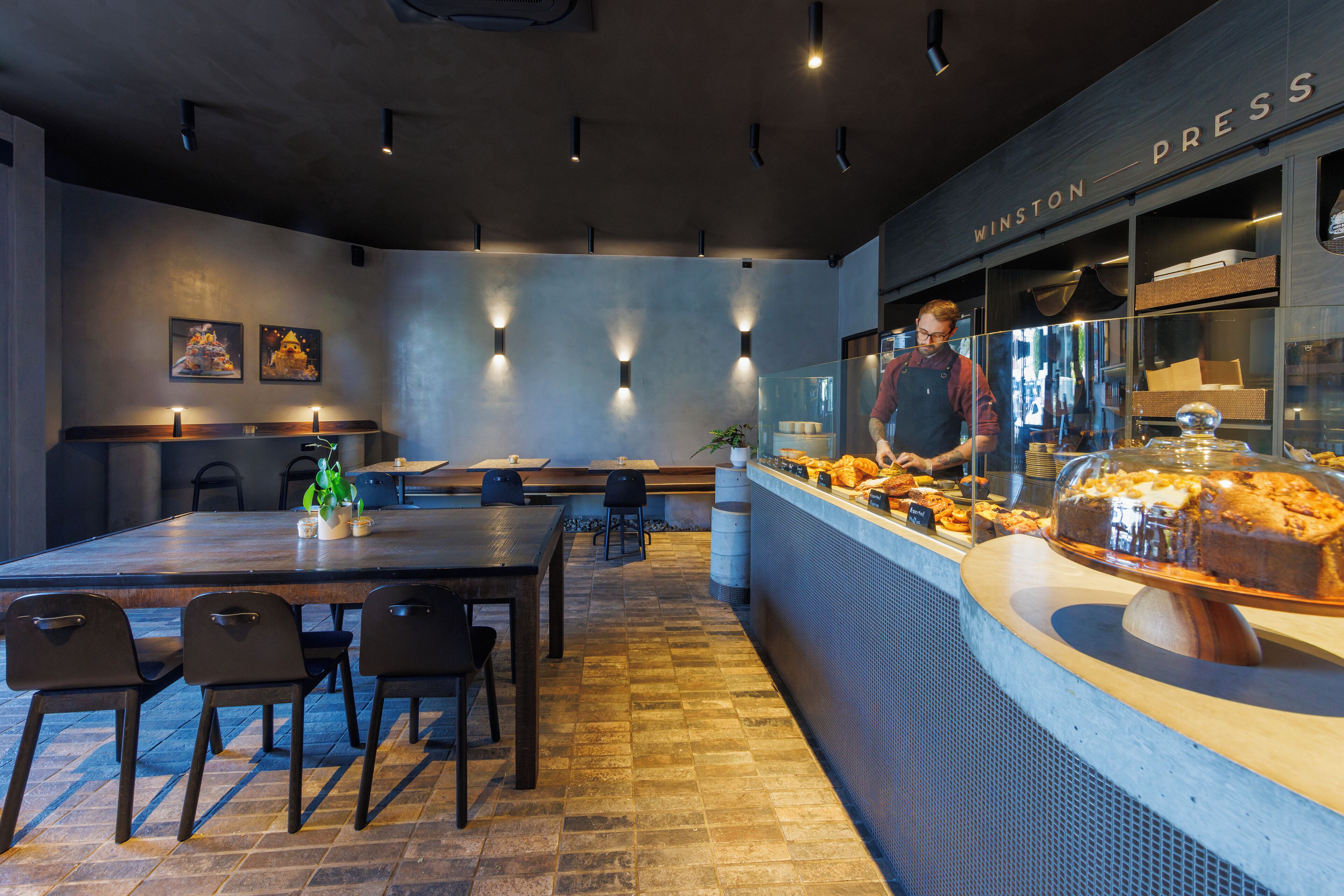L'intérieur d'un café moderne, avec une longue table en bois, des chaises noires, un comptoir où sont exposées des pâtisseries et une enseigne indiquant « WINSTON PRESS ».