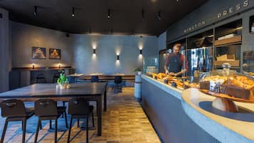 Ein modernes Café-Interieur mit einem langen Holztisch, schwarzen Stühlen, einer Theke mit Backwaren und einem 'WINSTON PRESS'-Schild.