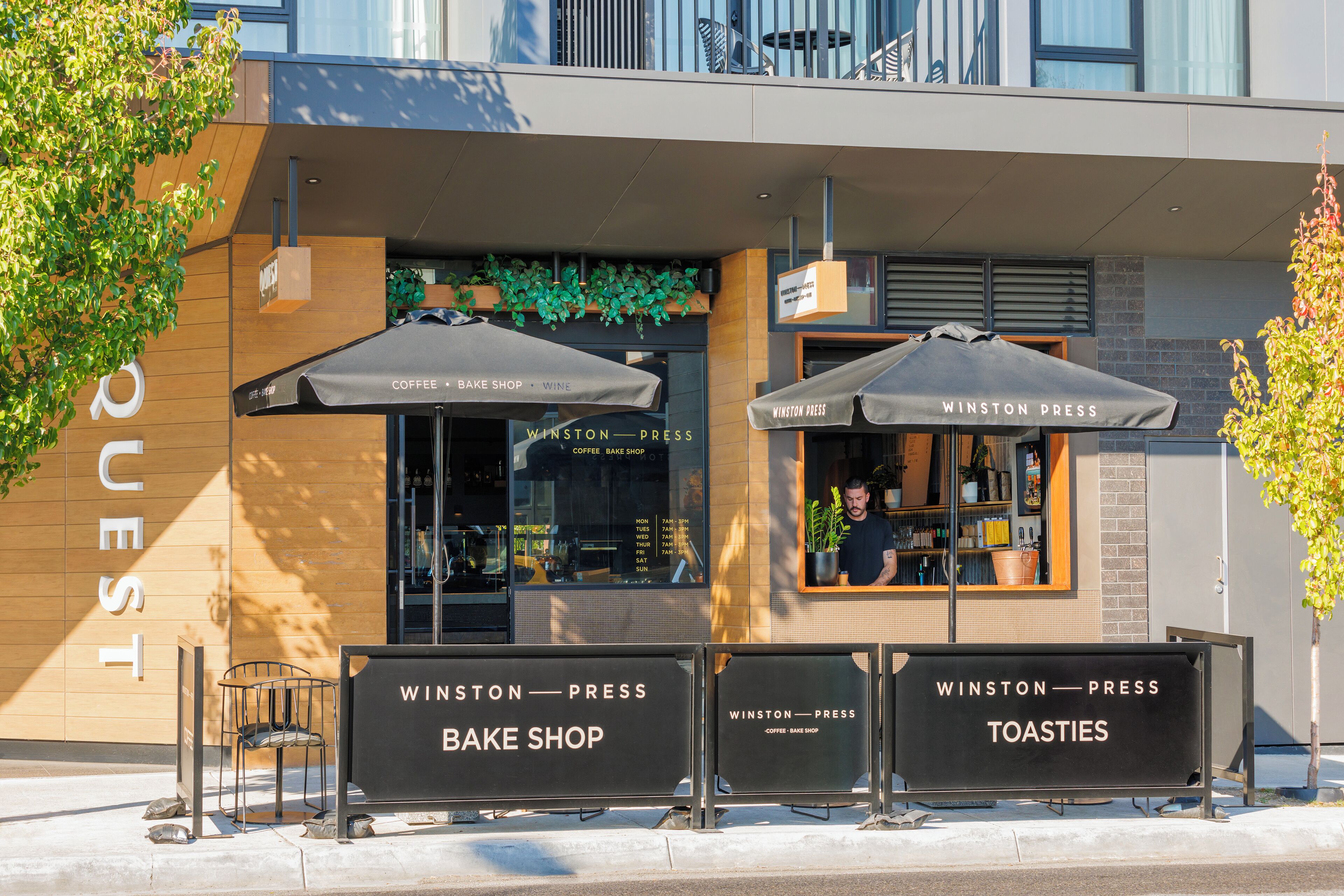 A modern building with a sign for 'Winston Press' cafe, featuring outdoor seating with umbrellas and a visible menu board.