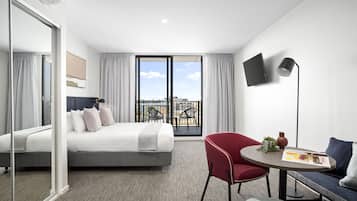 A modern hotel room with a bed, a red chair, a round table, a sofa, a TV, and a balcony with a city view.