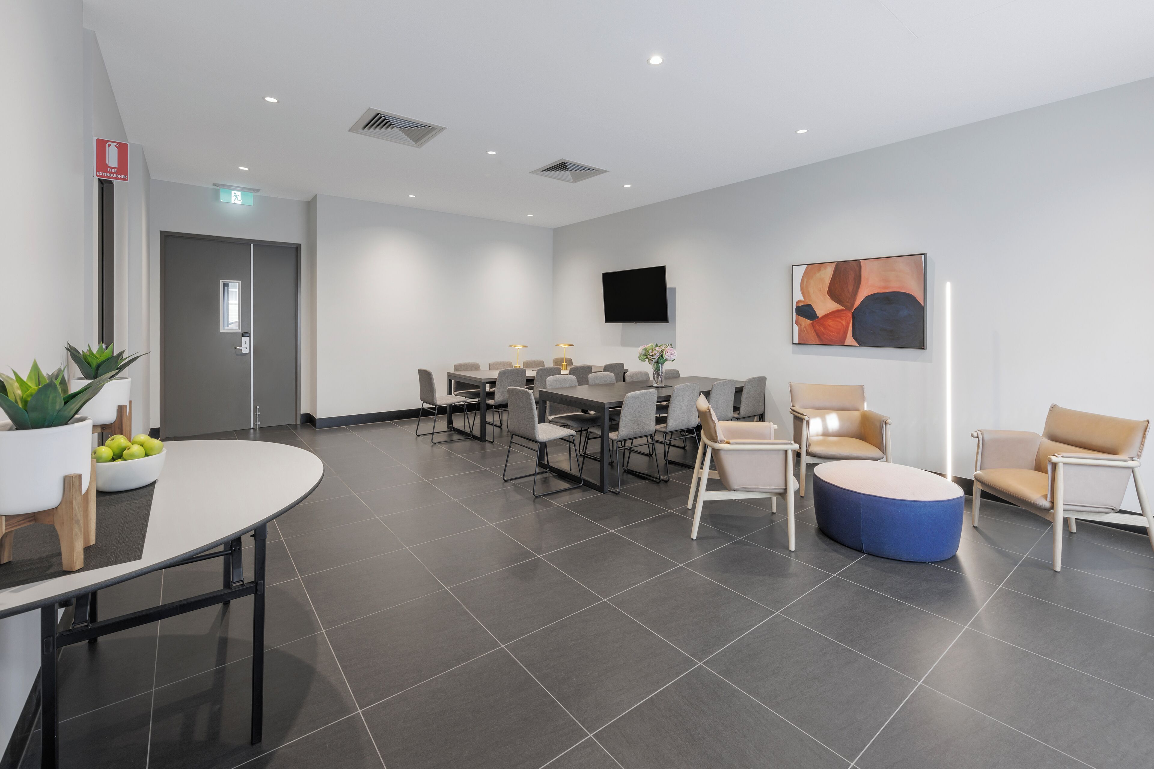 A modern conference room with a round table, several chairs, a blue ottoman, and a flat-screen TV on the wall.