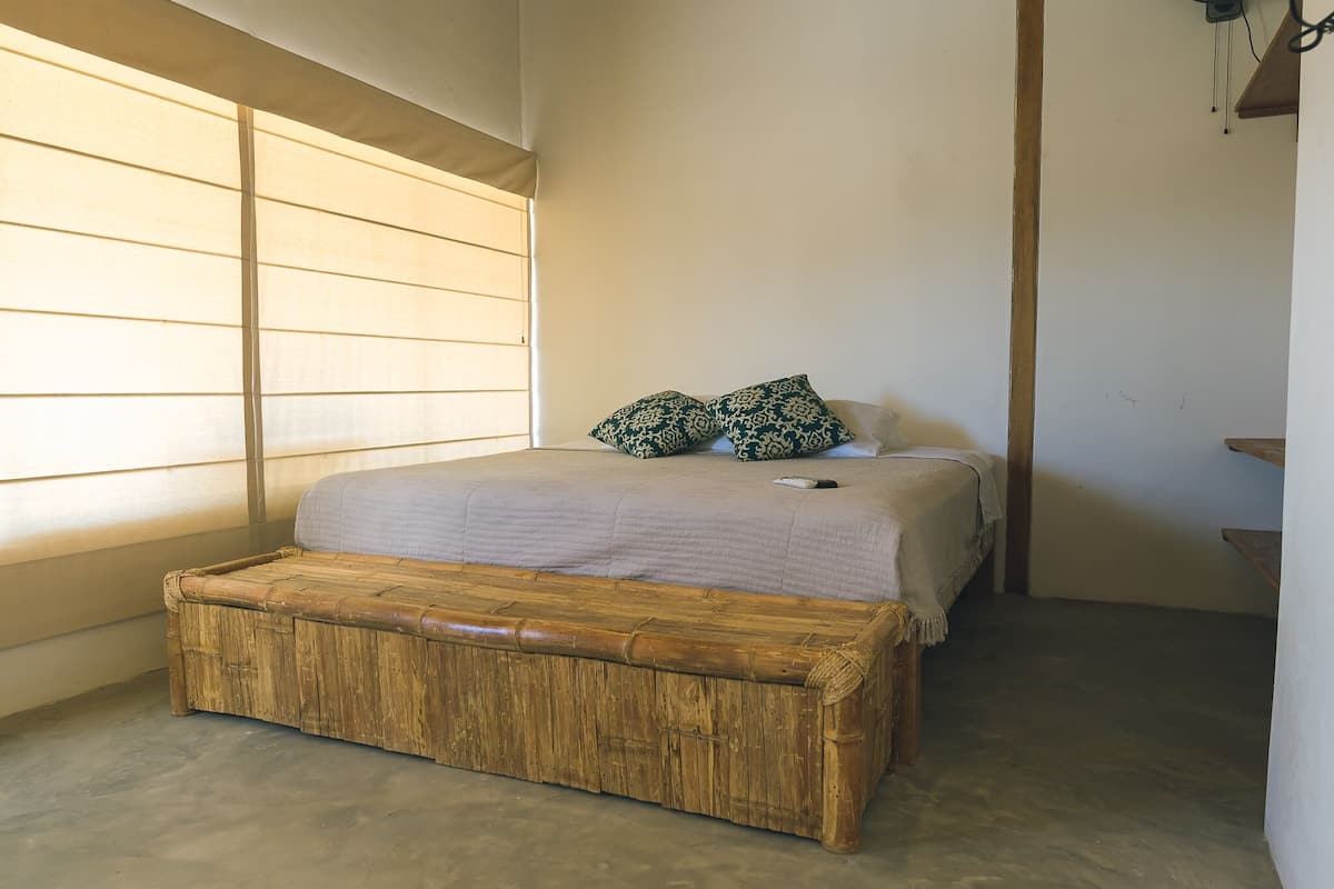 Une chambre à coucher avec un lit, deux coussins à motifs, un banc en bois et une fenêtre avec des stores.