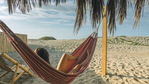 A hammock on a sandy beach with a wooden chair and a palm tree in the background. - Vistazul (Los Organos)