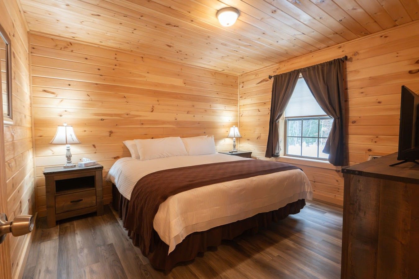Une chambre dans une cabane en bois, avec un lit, des tables de chevet, des lampes, une fenêtre avec des rideaux et un téléviseur posé sur une commode en bois.