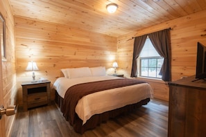Ein Schlafzimmer einer Holzhütte mit einem Bett, Nachttischen, Lampen, einem Fenster mit Vorhängen und einem Fernseher auf einer Holzkommode.